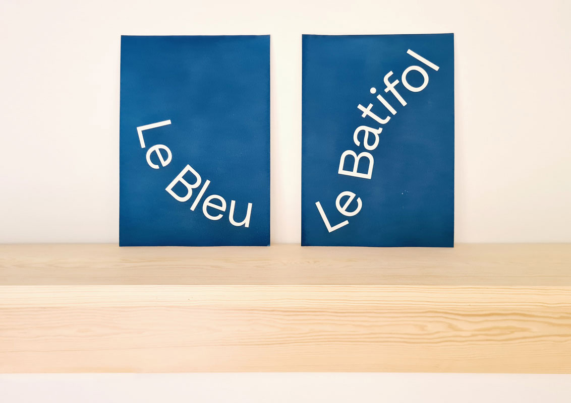 Deux affiches, &laquo;&nbsp;Le Bleu&nbsp;&raquo; et &laquo;&nbsp;Le Batifol&nbsp;&raquo;, c&ocirc;te &agrave; c&ocirc;te sur une &eacute;tag&egrave;re en bois.